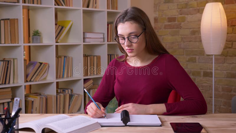 La giovane studentessa dai capelli lunghi castana in libro di lettura di vetro dell'occhio fa le note che sono attente in ufficio