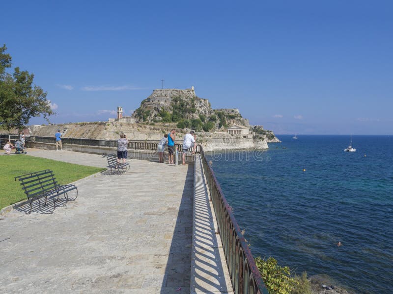 La Grecia, Corfù, città di Kerkyra, il 26 settembre 2018: Gente turistica su passeggiata con il banco, il parco e la vista su fam immagini stock La Grecia, Corfù, città di Kerkyra, il 26 settembre 2018: Gente turistica su passeggiata con il banco, il parco e la vista su fam immagini stock