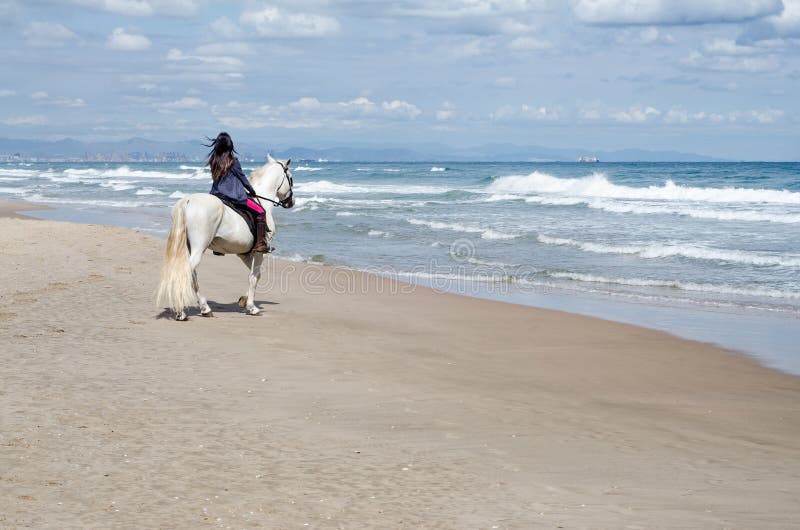 La guida della giovane donna lungo la spiaggia con il suo cavallo bianco, vede la città nei precedenti immagini stock libere da diritti La guida della giovane donna lungo la spiaggia con il suo cavallo bianco, vede la città nei precedenti immagini stock libere da diritti