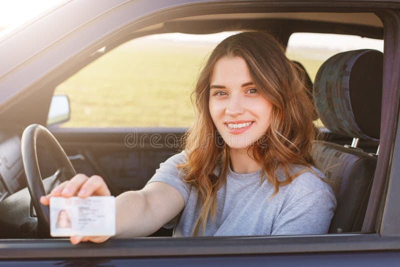 La hembra joven sonriente con aspecto agradable muestra orgulloso su licencia de conductores, se sienta en nuevo coche, siendo co fotos de archivo libres de regalías La hembra joven sonriente con aspecto agradable muestra orgulloso su licencia de conductores, se sienta en nuevo coche, siendo co fotos de archivo libres de regalías