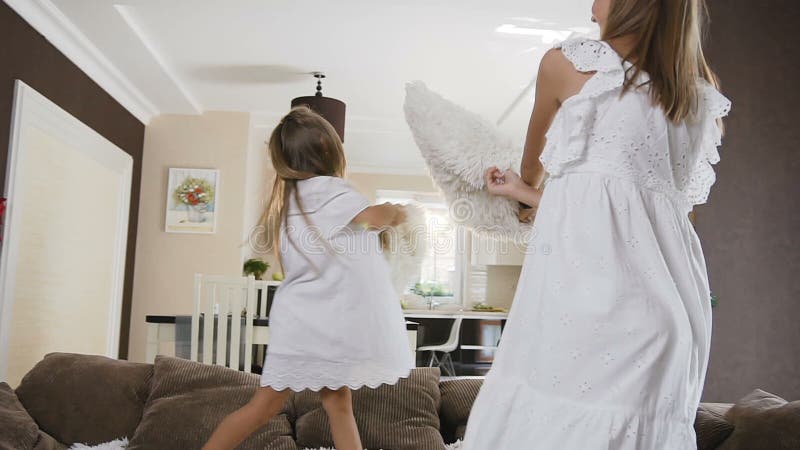 La hermana dos en el salto blanco del vestido en el sofá se divierte que lucha con las almohadas suaves en la sala de estar en ca almacen de video La hermana dos en el salto blanco del vestido en el sofá se divierte que lucha con las almohadas suaves en la sala de estar en ca almacen de video