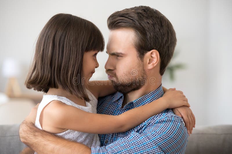 Diverse family father and adorable preschool daughter make funny faces blowing cheeks looking at each other have fun indoors. Profile male and little child with puffed cheeks sitting on couch at home. Diverse family father and adorable preschool daughter make funny faces blowing cheeks looking at each other have fun indoors. Profile male and little child with puffed cheeks sitting on couch at home