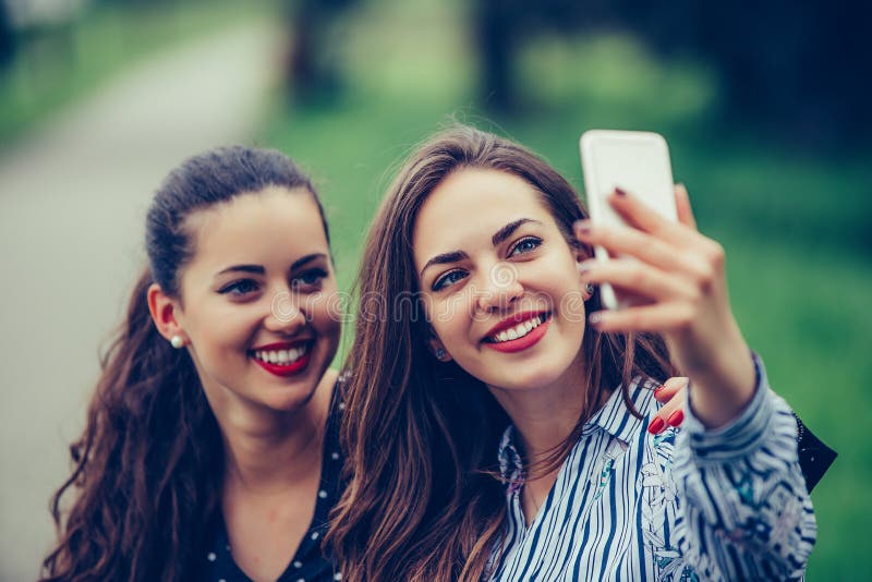 La imagen de los amigos hermosos jovenes de las mujeres, estudiantes que se sientan en el parque hace el selfie por el teléfono m imágenes de archivo libres de regalías La imagen de los amigos hermosos jovenes de las mujeres, estudiantes que se sientan en el parque hace el selfie por el teléfono m imágenes de archivo libres de regalías