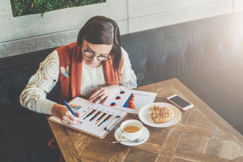 La jeune femme d'affaires dans les verres et le chandail blanc s'assied en café à la table, fonctionnant La femme regarde des dia images stock La jeune femme d'affaires dans les verres et le chandail blanc s'assied en café à la table, fonctionnant La femme regarde des dia images stock