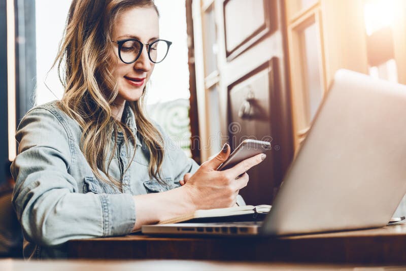 La jeune femme d'affaires s'assied dans le café à la table devant l'ordinateur et le carnet, utilisant le smartphone Medias socia image libre de droits La jeune femme d'affaires s'assied dans le café à la table devant l'ordinateur et le carnet, utilisant le smartphone Medias socia image libre de droits