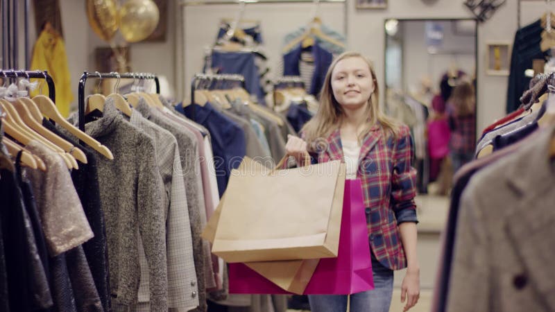 La jeune femme heureuse avec des sacs à provisions marche au support des vêtements dans un magasin et la recherche d'habillement