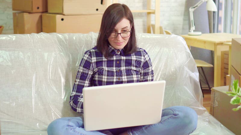 La jeune femme joyeuse de portrait utilise l'ordinateur portable se reposant sur le divan après déplacement à un appartement mode banque de vidéos