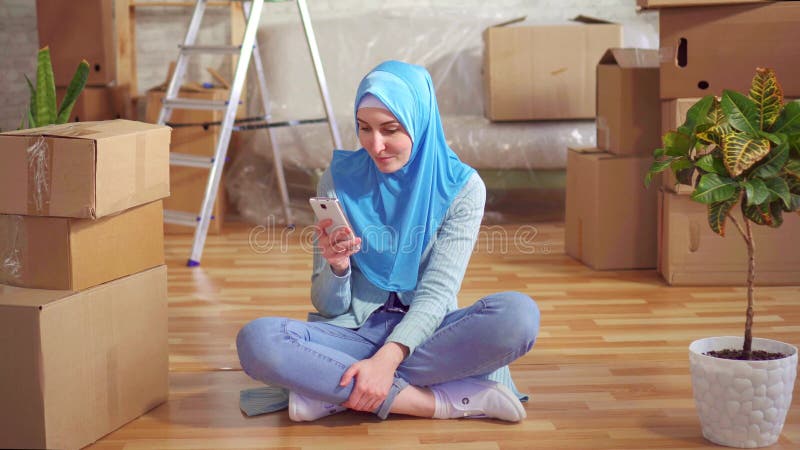 La jeune femme musulmane de portrait dans un hijab utilise le smartphone se reposant sur le plancher à côté des boîtes dans un ap banque de vidéos