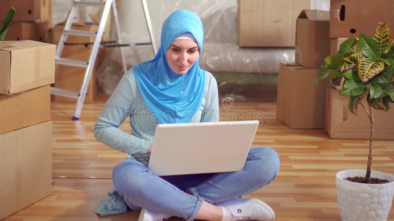 La jeune femme musulmane de portrait dans un hijab utilise un ordinateur portable se reposant sur le plancher à côté des boîtes clips vidéos