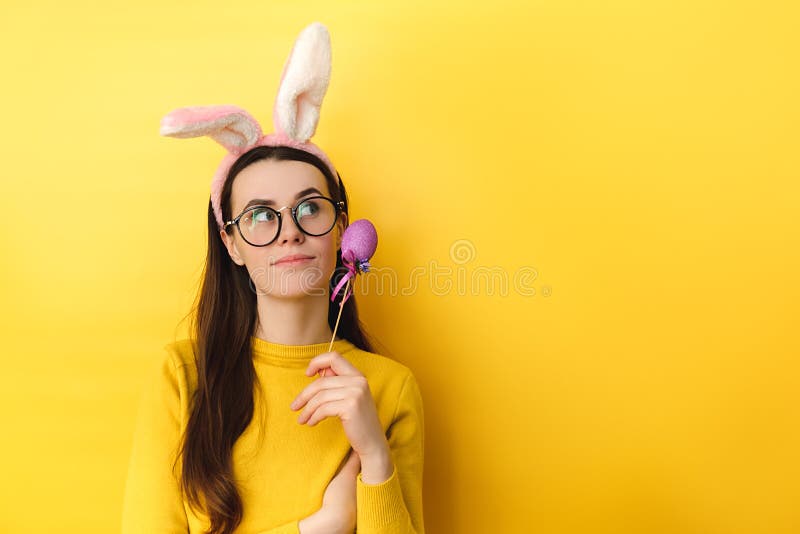 La jeune femme pensive en verres utilise les oreilles pelucheuses détient l'oeuf coloré et regarde contemple loin à propos de quel images stock