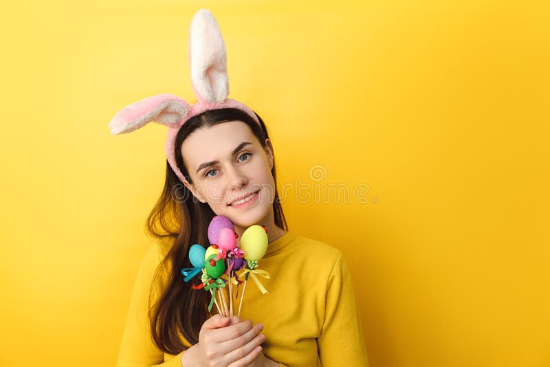 La jeune femme positive utilise les oreilles pelucheuses tient des oeufs colorés sur les bâtonnets étant dans la bonne humeur et a photos stock