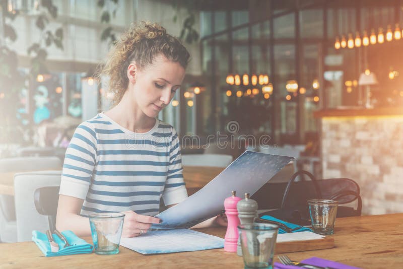 La jeune femme s'assied en café à la table en bois et à la lecture La fille attend les amis, collègues dans le restaurant images libres de droits La jeune femme s'assied en café à la table en bois et à la lecture La fille attend les amis, collègues dans le restaurant images libres de droits