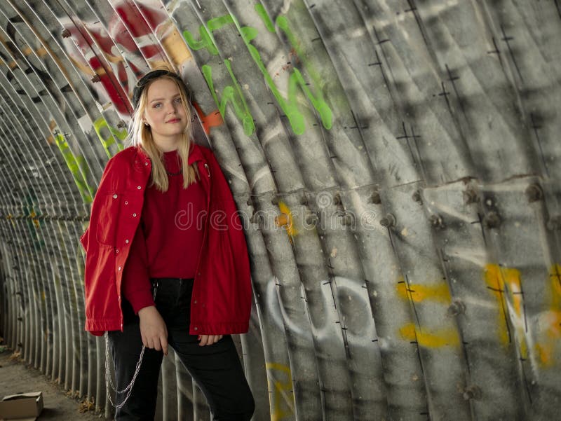 La jeune fille attirante de hippie dans le chapeau tricoté et la veste rouge se tient sous un pont dans la perspective d'un mur a image stock La jeune fille attirante de hippie dans le chapeau tricoté et la veste rouge se tient sous un pont dans la perspective d'un mur a image stock