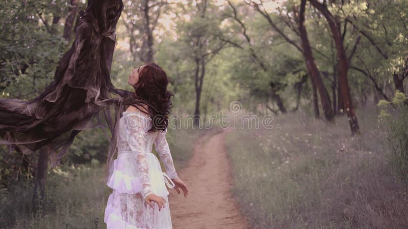 La jeune fille mignonne dans une longue robe transparente blanche avec un modèle floral et des cheveux onduleux foncés a été capt clips vidéos