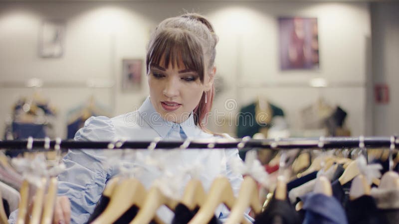 La jolie jeune femme marche par le support des vêtements dans un magasin d'habillement et prend à un examen rapide sur des vêteme