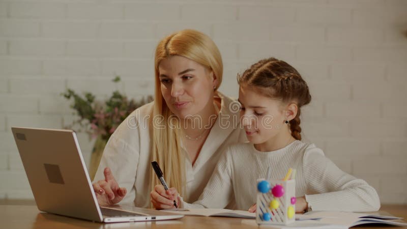 La joven madre ayuda a su hija con los deberes en su laptop. la linda madre y su hija hacen sus deberes. metrajes