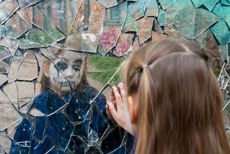 Young girl zombie looks in a broken mirror and shows her hand on a mirror. Halloween and day of the dead concept. Young girl zombie looks in a broken mirror and shows her hand on a mirror. Halloween and day of the dead concept.