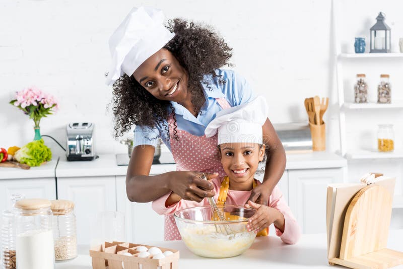 La madre afroamericana e la figlia in cappelli del cuoco unico che mescolano la pasta con sbattono fotografie stock