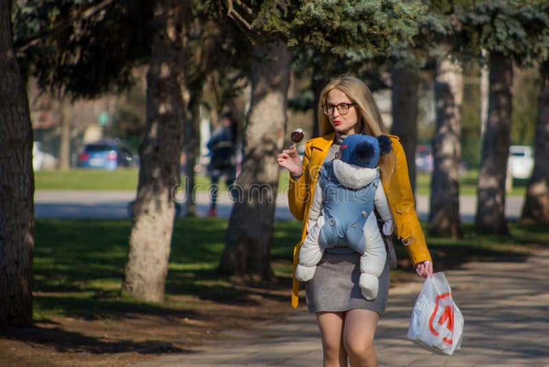 La madre linda en vidrios lleva al bebé en su pecho en correas de refrenamiento especiales y come el helado foto de archivo libre de regalías La madre linda en vidrios lleva al bebé en su pecho en correas de refrenamiento especiales y come el helado foto de archivo libre de regalías