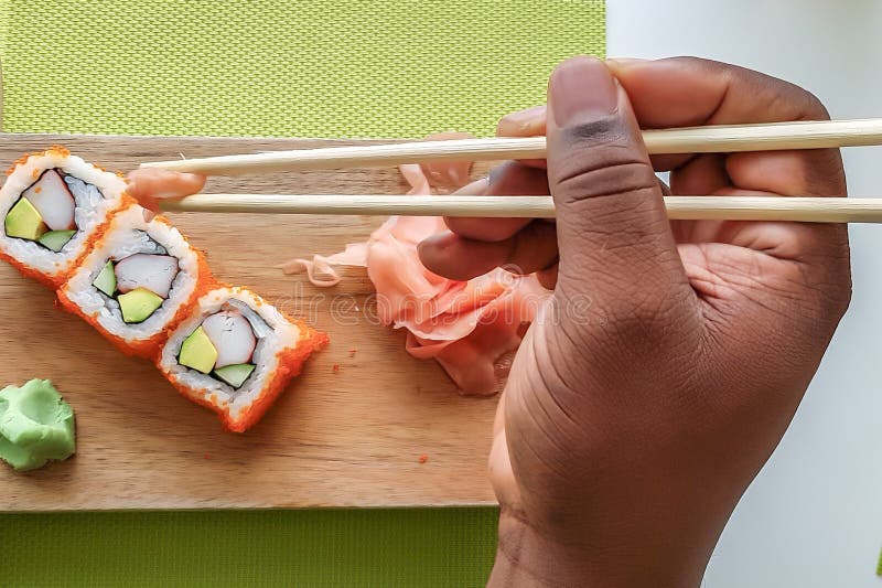 La mano del hombre que sostiene los palillos y que come los rollos de sushi de California en un tablero de madera imagen de archivo La mano del hombre que sostiene los palillos y que come los rollos de sushi de California en un tablero de madera imagen de archivo