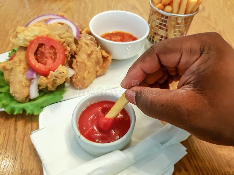 La mano della persona africana che tiene le patate fritte, immergenti in ketchup - mangiare alimento in un ristorante; Ali e sals fotografie stock libere da diritti La mano della persona africana che tiene le patate fritte, immergenti in ketchup - mangiare alimento in un ristorante; Ali e sals fotografie stock libere da diritti