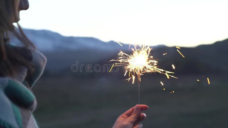 La mano femenina sostiene la bengala en un fondo de montañas nevosas, puesta del sol hermosa