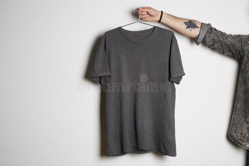 La mano maschio tatuata tiene la caduta con la maglietta grigia in bianco dal premio leggermente capisce il fondo bianco Pasta de fotografie stock La mano maschio tatuata tiene la caduta con la maglietta grigia in bianco dal premio leggermente capisce il fondo bianco Pasta de fotografie stock