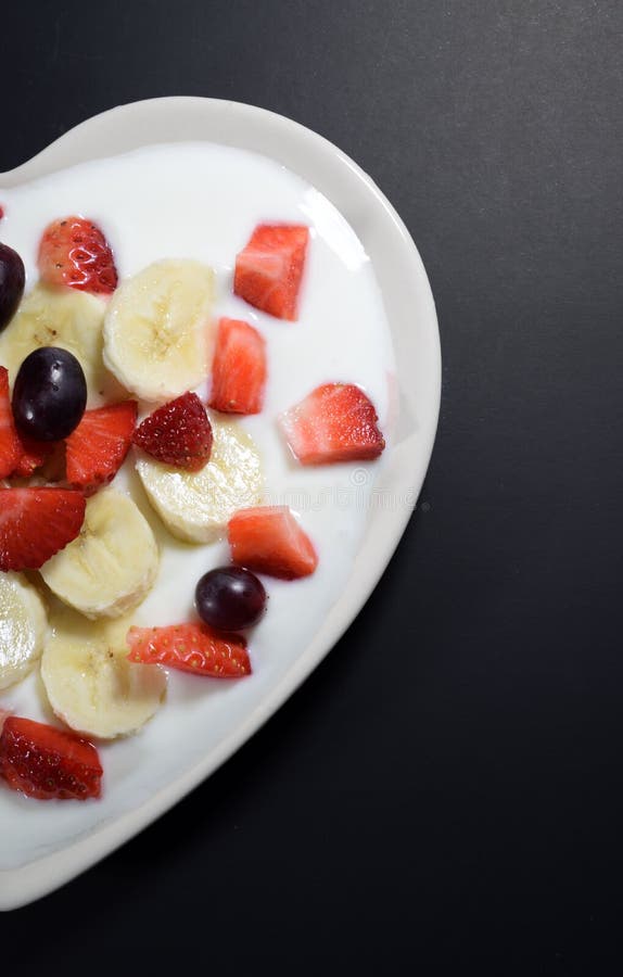 La metà del piatto crema in forma di cuore riempito di yogurt con le fette della banana, fragole incide i pezzi e le bacche dell immagini stock libere da diritti La metà del piatto crema in forma di cuore riempito di yogurt con le fette della banana, fragole incide i pezzi e le bacche dell immagini stock libere da diritti
