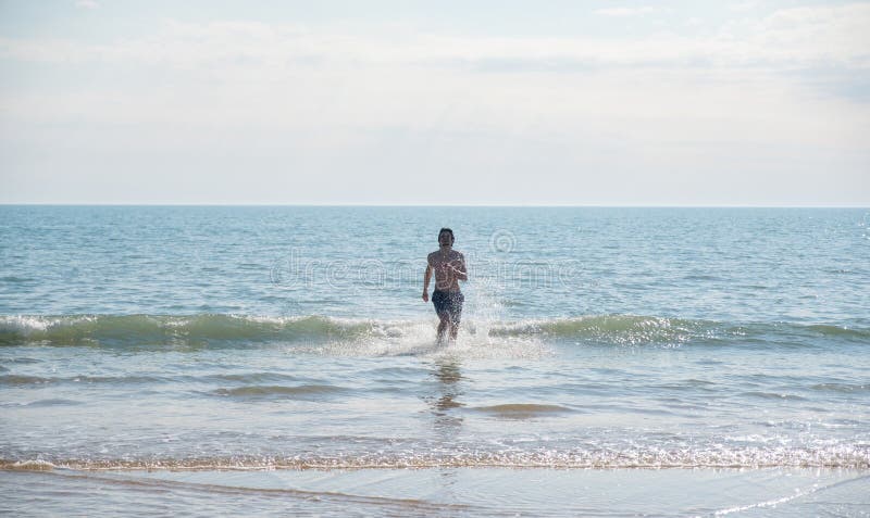 La morenita irreconocible del hombre en pantalones cortos azules corre fuera del mar en el verano, playa arenosa fotografía de archivo La morenita irreconocible del hombre en pantalones cortos azules corre fuera del mar en el verano, playa arenosa fotografía de archivo
