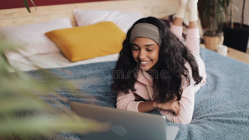 La muchacha afroamericana feliz miente en su vientre en la cama por la ventana que charla feliz con sus amigos en una llamada vid