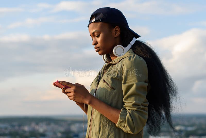 La muchacha afroamericana hermosa escucha la música y goza Mujer negra joven sonriente en fondo borroso de la ciudad foto de archivo La muchacha afroamericana hermosa escucha la música y goza Mujer negra joven sonriente en fondo borroso de la ciudad foto de archivo