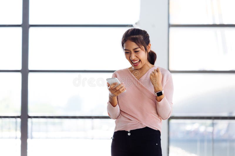 La muchacha asiática joven que mira su teléfono móvil y consigue excitada dentro del edificio fotos de archivo La muchacha asiática joven que mira su teléfono móvil y consigue excitada dentro del edificio fotos de archivo