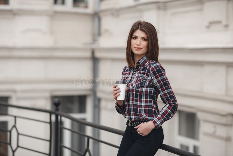 La muchacha bonita, morenita hermosa con el peinado elegante en una camisa de tela escocesa bebe la taza de café al aire libre en fotografía de archivo La muchacha bonita, morenita hermosa con el peinado elegante en una camisa de tela escocesa bebe la taza de café al aire libre en fotografía de archivo