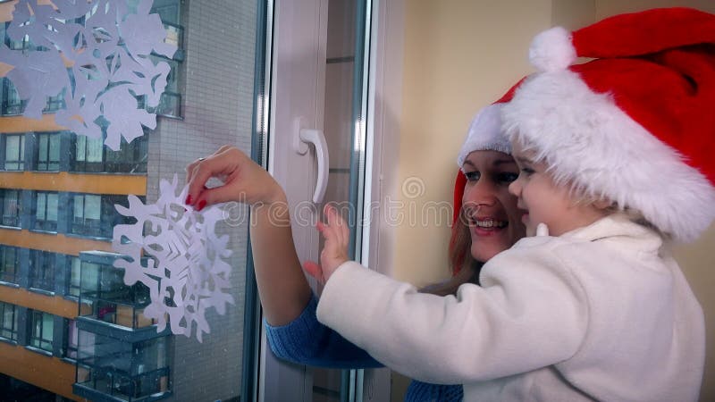 La muchacha cariñosa de la mujer y del niño con los sombreros rojos de santa pega los copos de nieve de papel en ventana