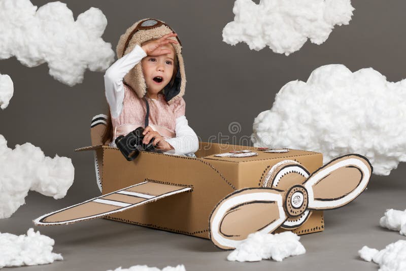 La muchacha del niño juega en un aeroplano hecho de la caja de cartón y de sueños de hacer piloto, nubes de la algodón en un back imagen de archivo La muchacha del niño juega en un aeroplano hecho de la caja de cartón y de sueños de hacer piloto, nubes de la algodón en un back imagen de archivo