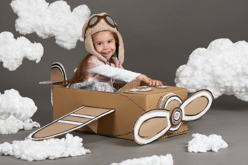 La muchacha del niño juega en un aeroplano hecho de la caja de cartón y de sueños de hacer piloto, nubes de la algodón en un back imagen de archivo libre de regalías La muchacha del niño juega en un aeroplano hecho de la caja de cartón y de sueños de hacer piloto, nubes de la algodón en un back imagen de archivo libre de regalías