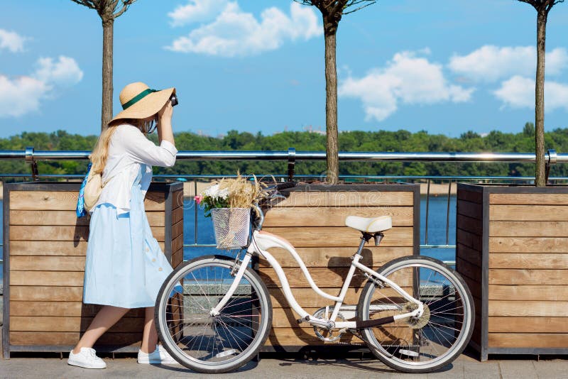 La muchacha en vestido azul y el sombrero de moda toman una imagen cerca de la bicicleta foto de archivo La muchacha en vestido azul y el sombrero de moda toman una imagen cerca de la bicicleta foto de archivo