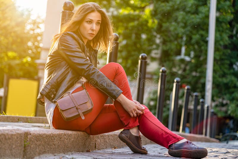 La muchacha está planteando la sentada atractiva en el encintado en pantalones rojos, chaqueta negra al aire libre Fotografía de imagen de archivo libre de regalías La muchacha está planteando la sentada atractiva en el encintado en pantalones rojos, chaqueta negra al aire libre Fotografía de imagen de archivo libre de regalías
