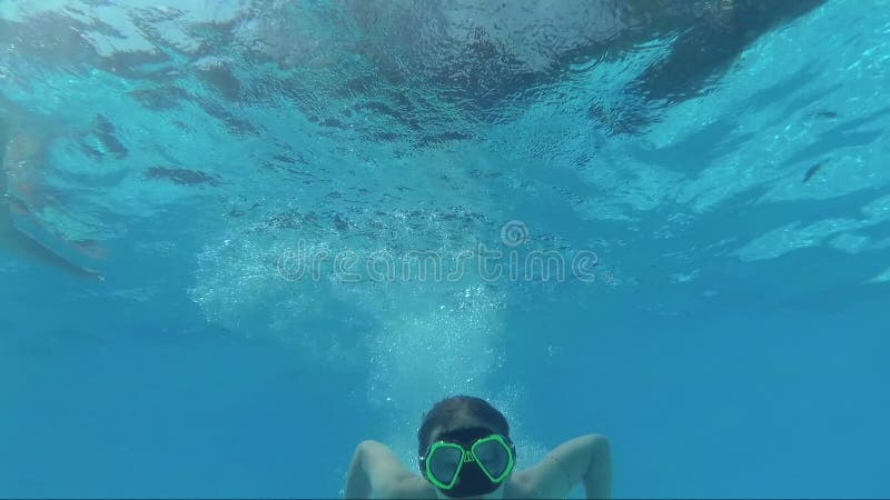 La muchacha feliz en una máscara debajo del agua en la piscina mira en la cámara