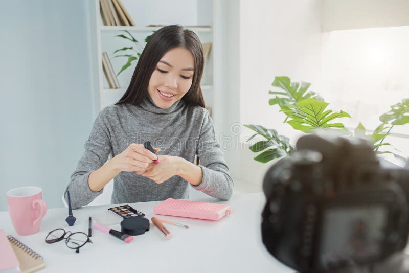 La muchacha hermosa se está sentando en la tabla y está haciendo un vídeo sobre sus productos de belleza del mes Ella está sosten fotos de archivo La muchacha hermosa se está sentando en la tabla y está haciendo un vídeo sobre sus productos de belleza del mes Ella está sosten fotos de archivo
