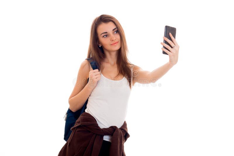 La muchacha joven alegre del estudiante con la mochila hace el selfie en su teléfono móvil aislado en el fondo blanco años del es foto de archivo La muchacha joven alegre del estudiante con la mochila hace el selfie en su teléfono móvil aislado en el fondo blanco años del es foto de archivo