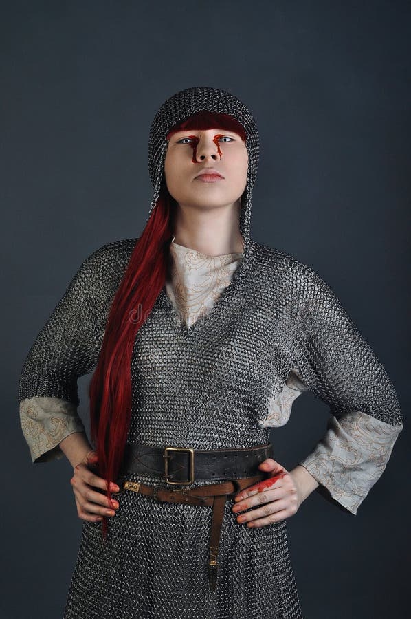 La muchacha medieval en correo en cadena con el pelo rojo se coloca en un fondo gris con sangre en sus manos fotografía de archivo libre de regalías La muchacha medieval en correo en cadena con el pelo rojo se coloca en un fondo gris con sangre en sus manos fotografía de archivo libre de regalías
