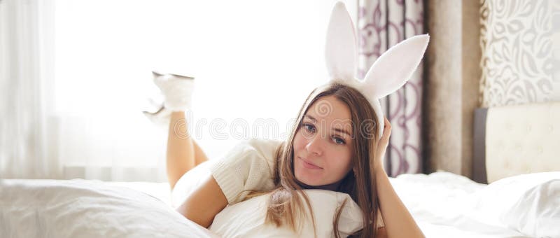 La muchacha morena preciosa con los oídos del conejito en su cabeza está mintiendo en una cama en su dormitorio foto de archivo libre de regalías La muchacha morena preciosa con los oídos del conejito en su cabeza está mintiendo en una cama en su dormitorio foto de archivo libre de regalías