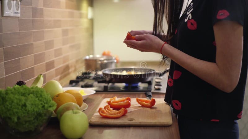 La muchacha negro-cabelluda linda en pijamas está cocinando las verduras para el desayuno en casa Ella es de limpieza y que corta almacen de metraje de vídeo