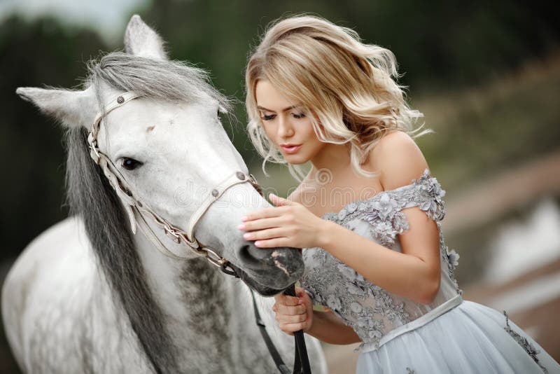 Beautiful blonde girl in dress strokes a gray horse on nature in summer. Beautiful blonde girl in dress strokes a gray horse on nature in summer