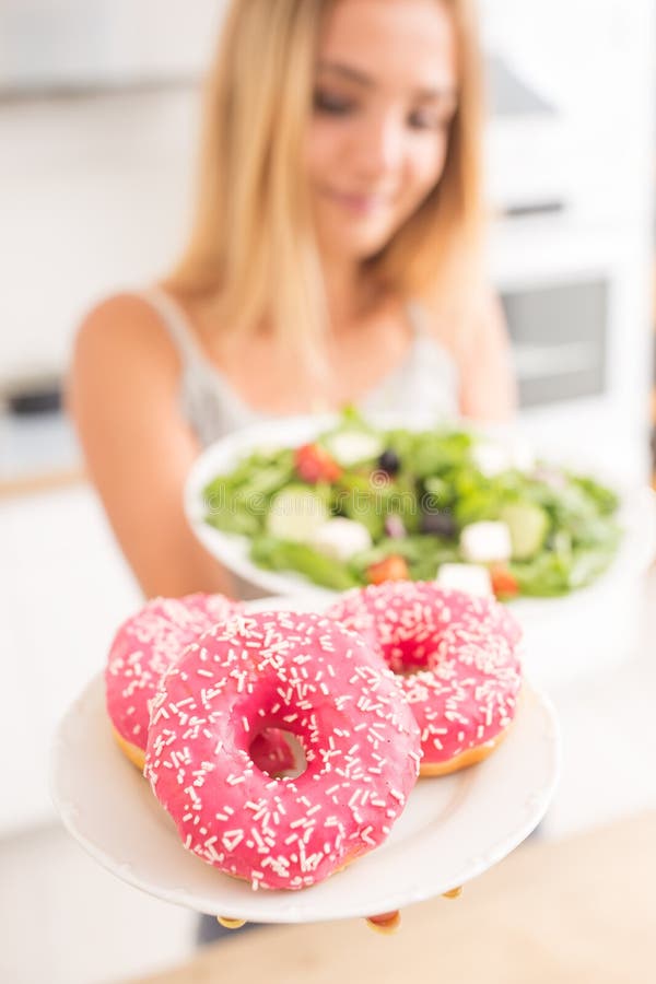 La muchacha rubia joven en el desayuno o la cena en la cocina casera elige entre el buñuelo y la ensalada vegetal imágenes de archivo libres de regalías La muchacha rubia joven en el desayuno o la cena en la cocina casera elige entre el buñuelo y la ensalada vegetal imágenes de archivo libres de regalías
