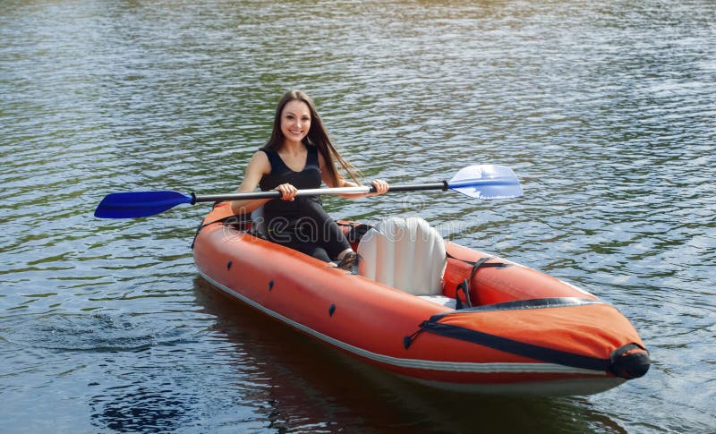 La muchacha sonriente - la deportista con el pelo del longdark en blacksportswear rema con un remo en el lago en una canoa inflab
