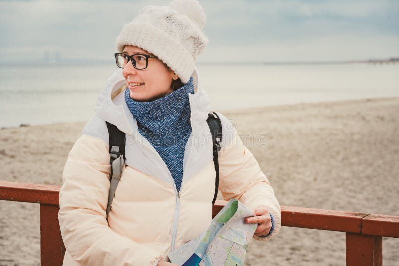 La mujer caucásica en sombrero y chaqueta con la mochila en invierno se sienta en el embarcadero de madera en la playa cerca de M fotos de archivo La mujer caucásica en sombrero y chaqueta con la mochila en invierno se sienta en el embarcadero de madera en la playa cerca de M fotos de archivo