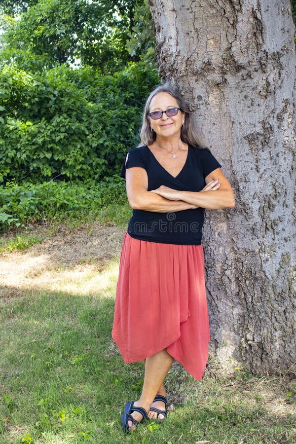 La mujer con el pelo y los vidrios grises largos y el organge bordean y la blusa del negro se inclina contra árbol grande con los fotografía de archivo La mujer con el pelo y los vidrios grises largos y el organge bordean y la blusa del negro se inclina contra árbol grande con los fotografía de archivo