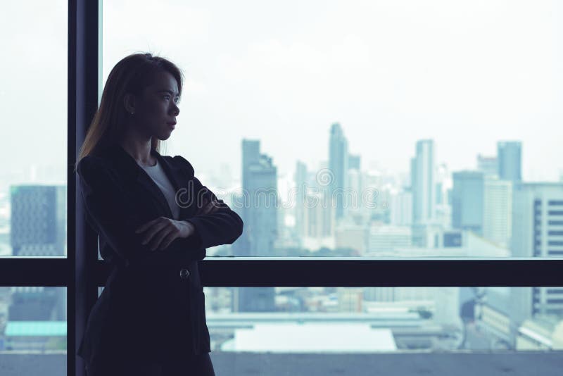 La mujer de negocios asi?tica cruz? los brazos que miraban hacia fuera la ventana el fondo de la opini?n de la ciudad imagen de archivo libre de regalías La mujer de negocios asi?tica cruz? los brazos que miraban hacia fuera la ventana el fondo de la opini?n de la ciudad imagen de archivo libre de regalías